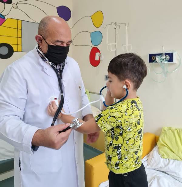 Dr. Sahib Cəfərov uşağın ağciyərlərini dinləyir, uşaq da stetoskopu qulağına taxıb